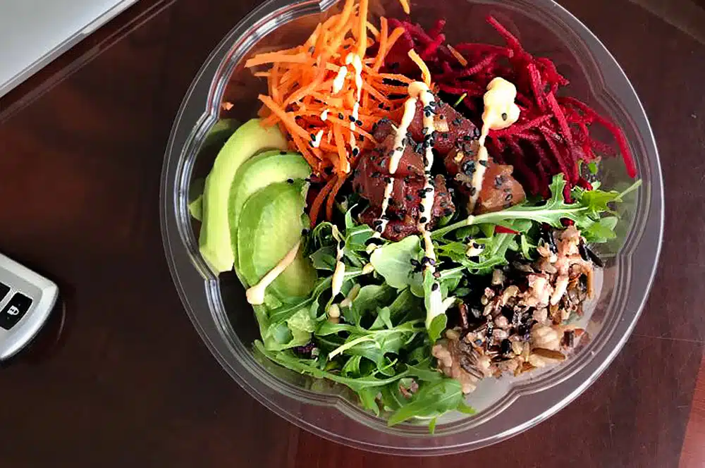 A close-up of an assembled poke bowl in a plastic salad bowl on a table with car keys and a MacBook.