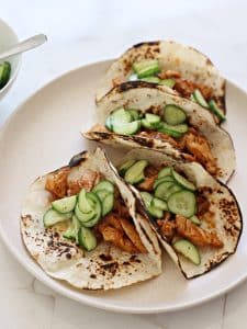 4 charred wheat wraps, each filled with chicken strips and seasoned cucumbers, on a plate.