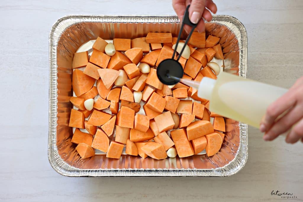 Filling a tbsp with oil over a 9x13 pan with sweet potato chunks and whole garlic cloves.