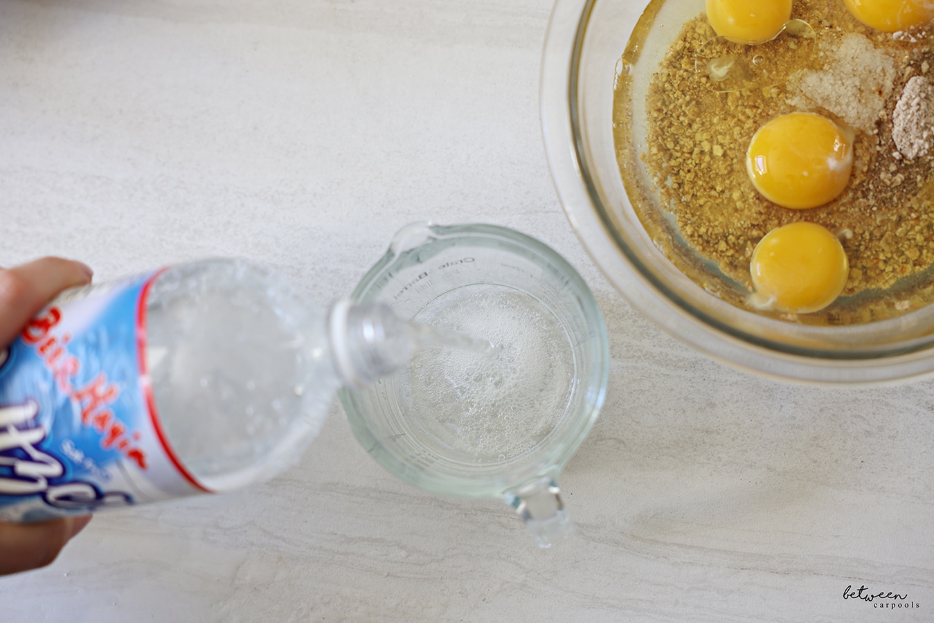 Pouring ¼ cup Be'er Mayim Seltzer into a glass measuring cup. Glass mixing bowl with Matzah Ball ingredients (eggs, oil, and matzah meal) nearby.