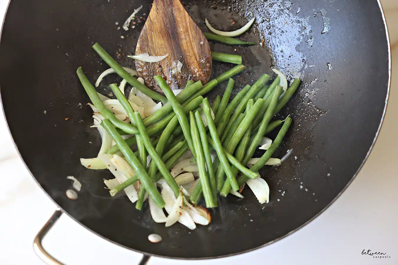 Green beans added to a Frenched onion and minced garlic sautéing in a wok with a wooden spoon.