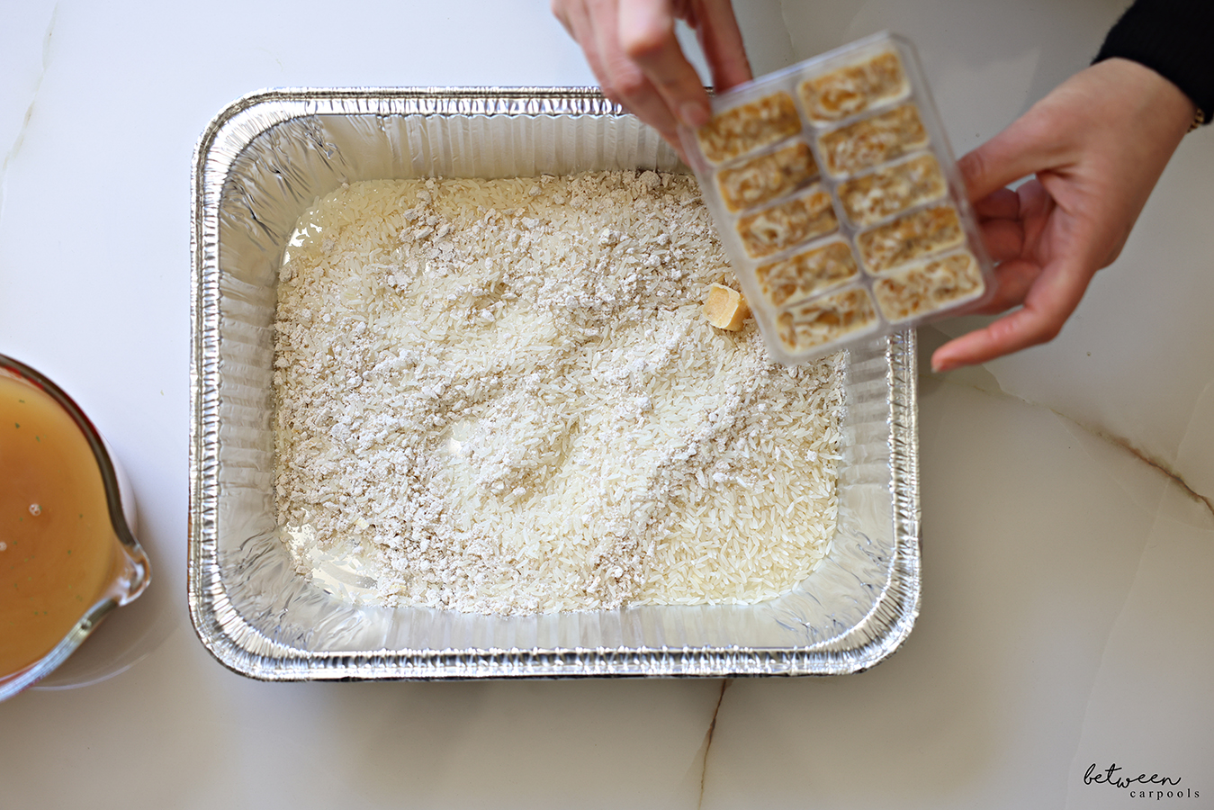 Adding frozen onion cubes to a 9x13 baking pan with jasmine rice, onion soup mix, and frozen garlic cubes.