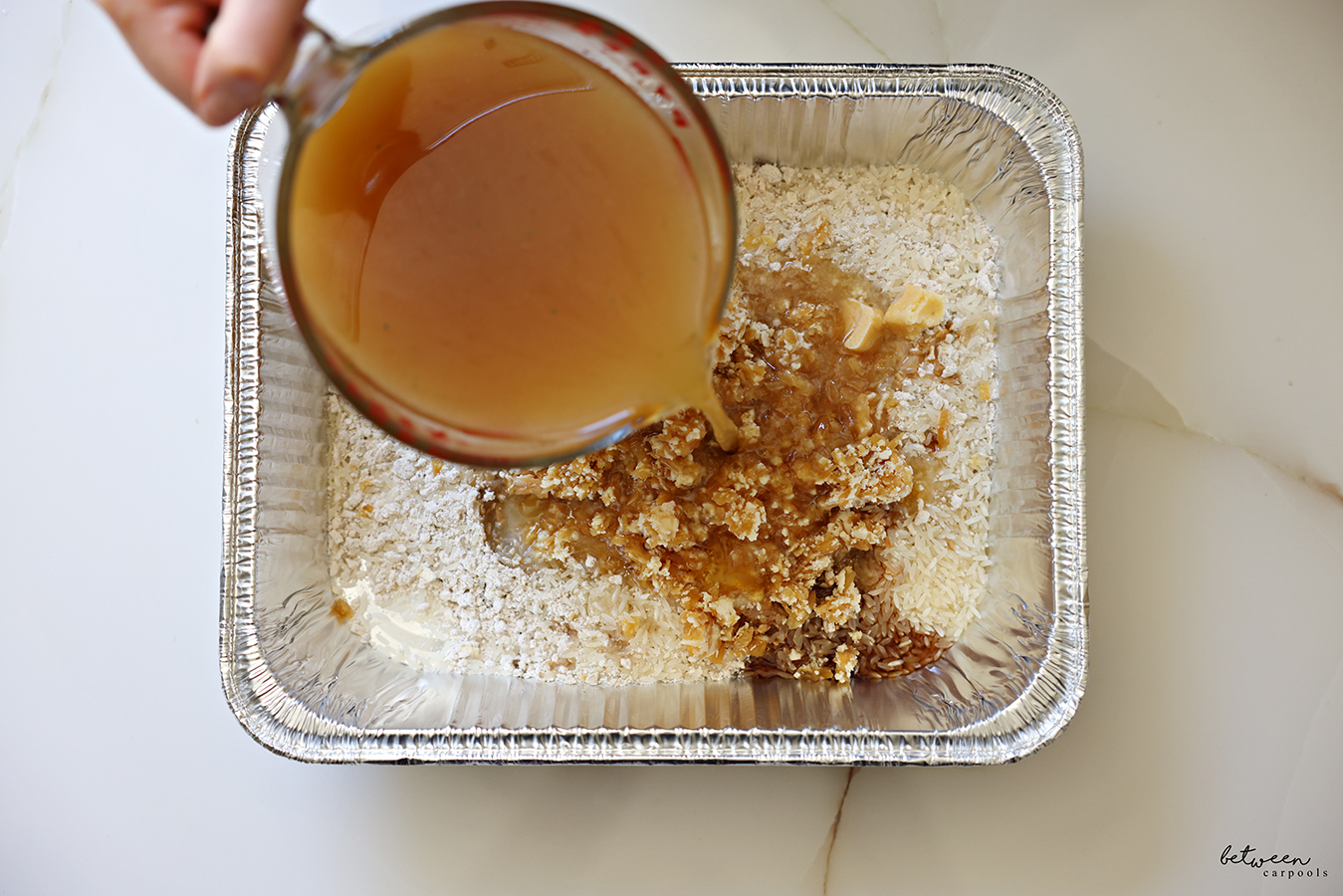 Pouring hot beef broth over jasmine rice, onion soup mix, frozen garlic and onion cubes in a 9x13 baking pan.