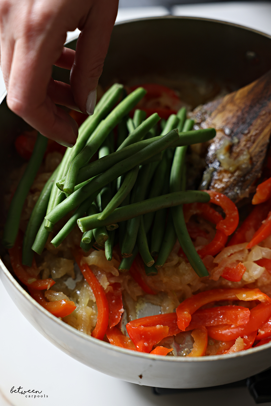 Adding French green beans to sautéed onion, garlic, and red pepper slices in a sauté pan.