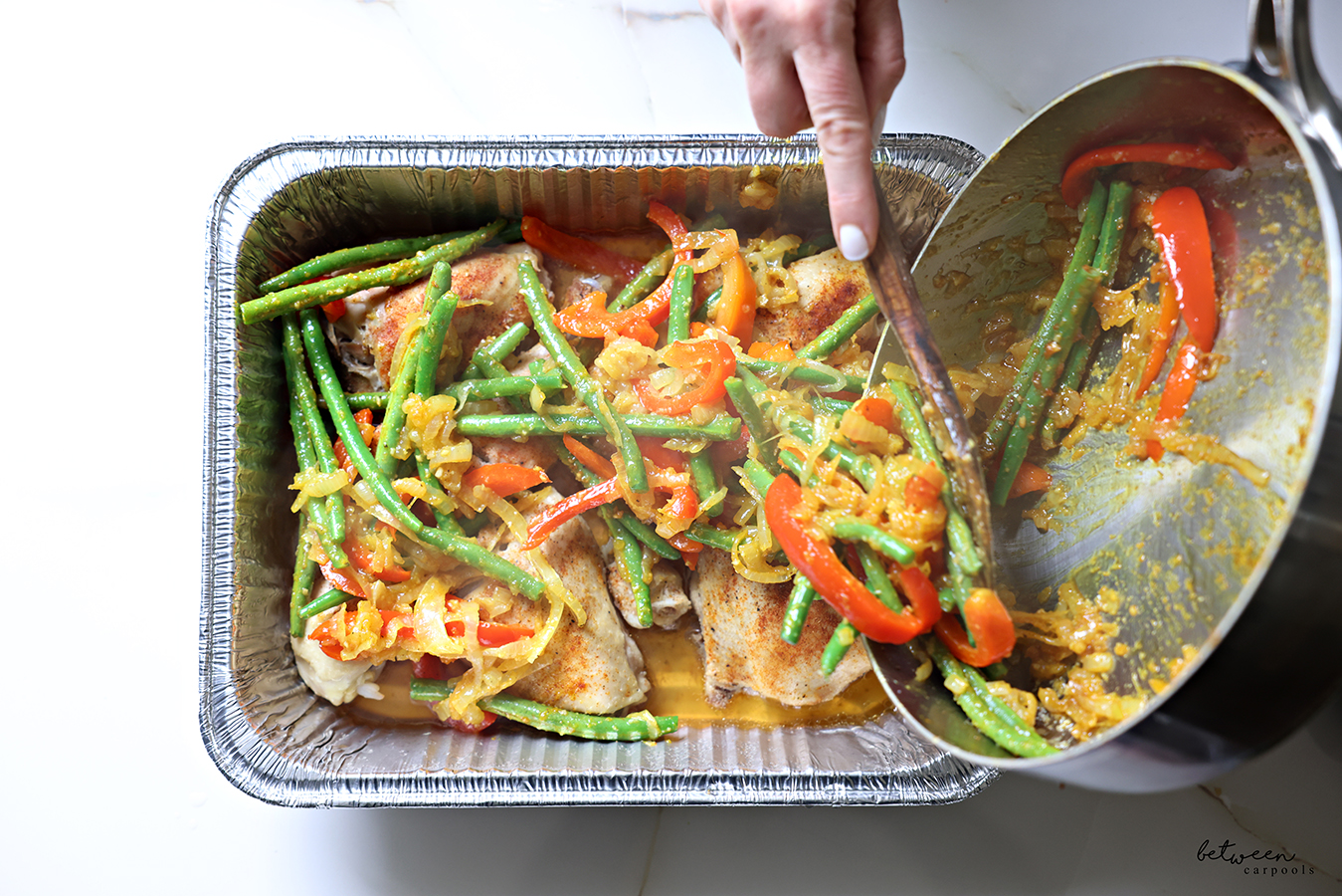 Topping baked chicken in a 9 x 13 aluminum pan with sautéed vegetables from a sauté pan, with a wooden spoon.