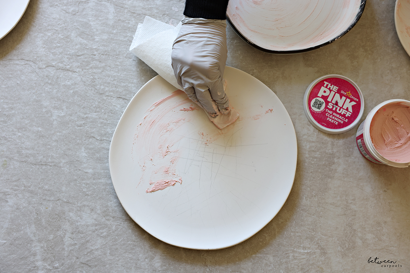 Scrubbing a matte white dinner plate with gloved hands and a slightly damp paper towel with The Pink Stuff.
