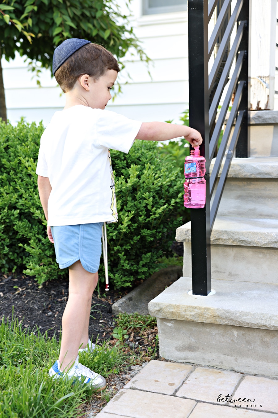 A little boy dipping a bubble stick into a bottle of Bubble Fun zip-tied to an outdoor stairs newel post.