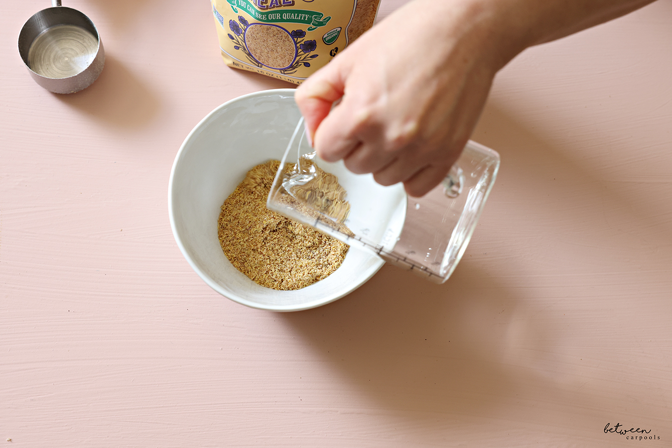 Adding water to flaxseed meal in a small bowl. Bag of flaxseed meal and a stainless measuring cup nearby.