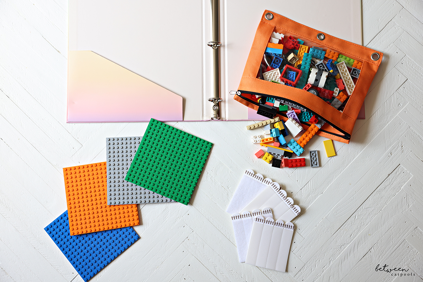 An open hardcover binder, a zippered pencil pouch with Lego bricks, some Lego plates and picture hanging strips.