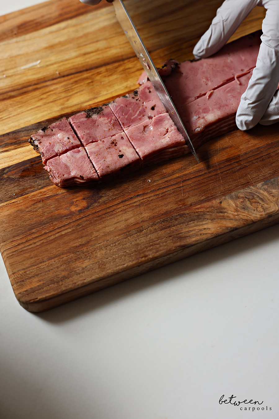 Cutting deli pastrami into cubes on a wooden cutting board with gloved hands and a knife.