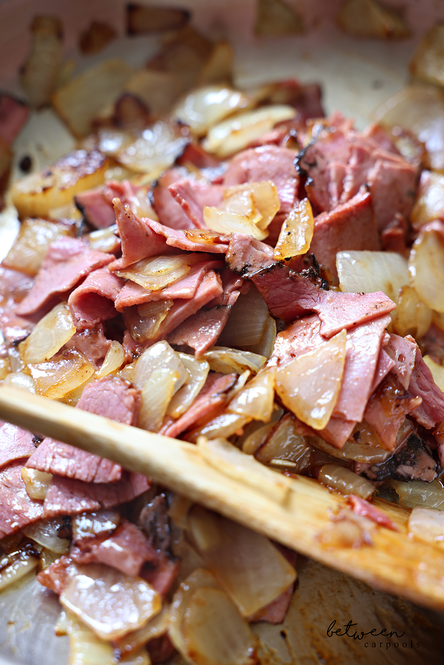 Closeup of sautéed onion chunks and deli pastrami cubes with BBQ sauce in a sauté pan with a wooden paddle.