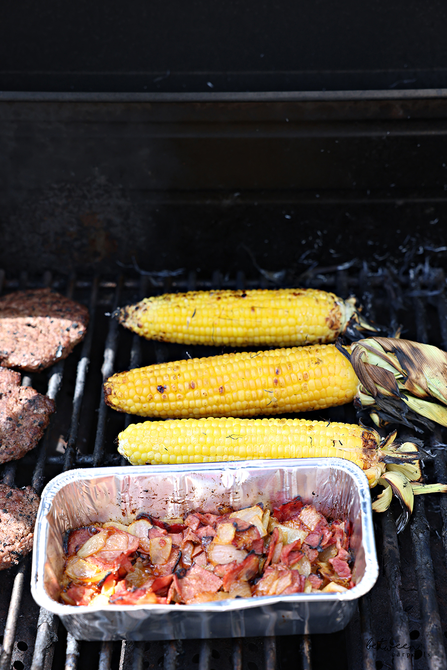 Aluminum loaf pan with onion pastrami topping on a grill with corn on the cob and burgers.