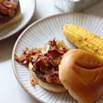 Burger topped with pastrami topping in a bun on a plate with corn on the cob on the side.