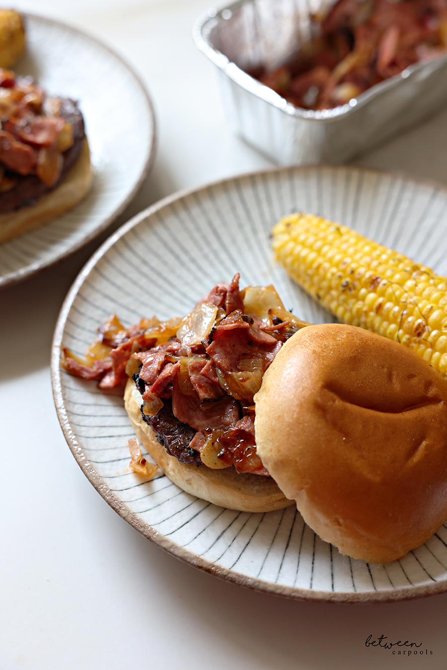 Burger topped with pastrami topping in a bun on a plate with corn on the cob on the side.