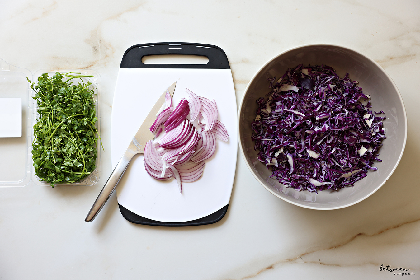 Bunch of parsley leaves, thinly sliced red onion on a plastic cutting board with a hast knife, and a salad bowl with shredded red cabbage.