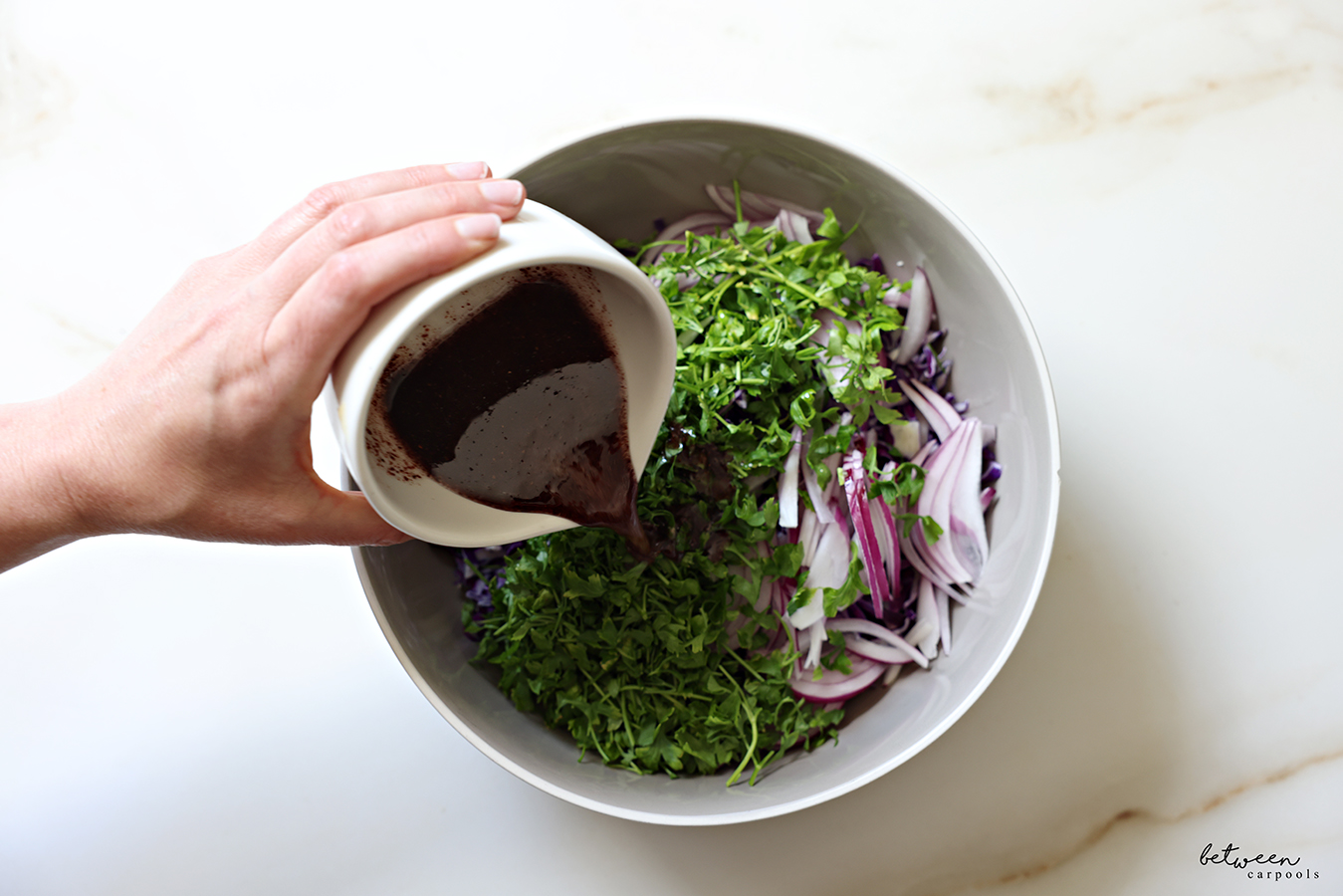 Pouring salad dressing over cabbage, red onion, and parsley in a large bowl.