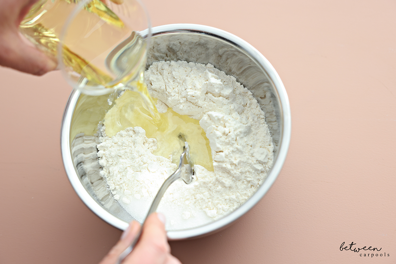 Pouring half a cup oil into a metal mixing bowl with flour and sugar.
