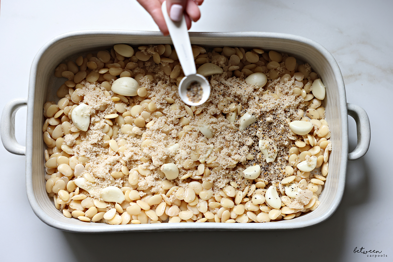 Adding Montreal steak seasoning to a roasting pan with boiled lima beans, whole garlic cloves, salt and brown sugar.