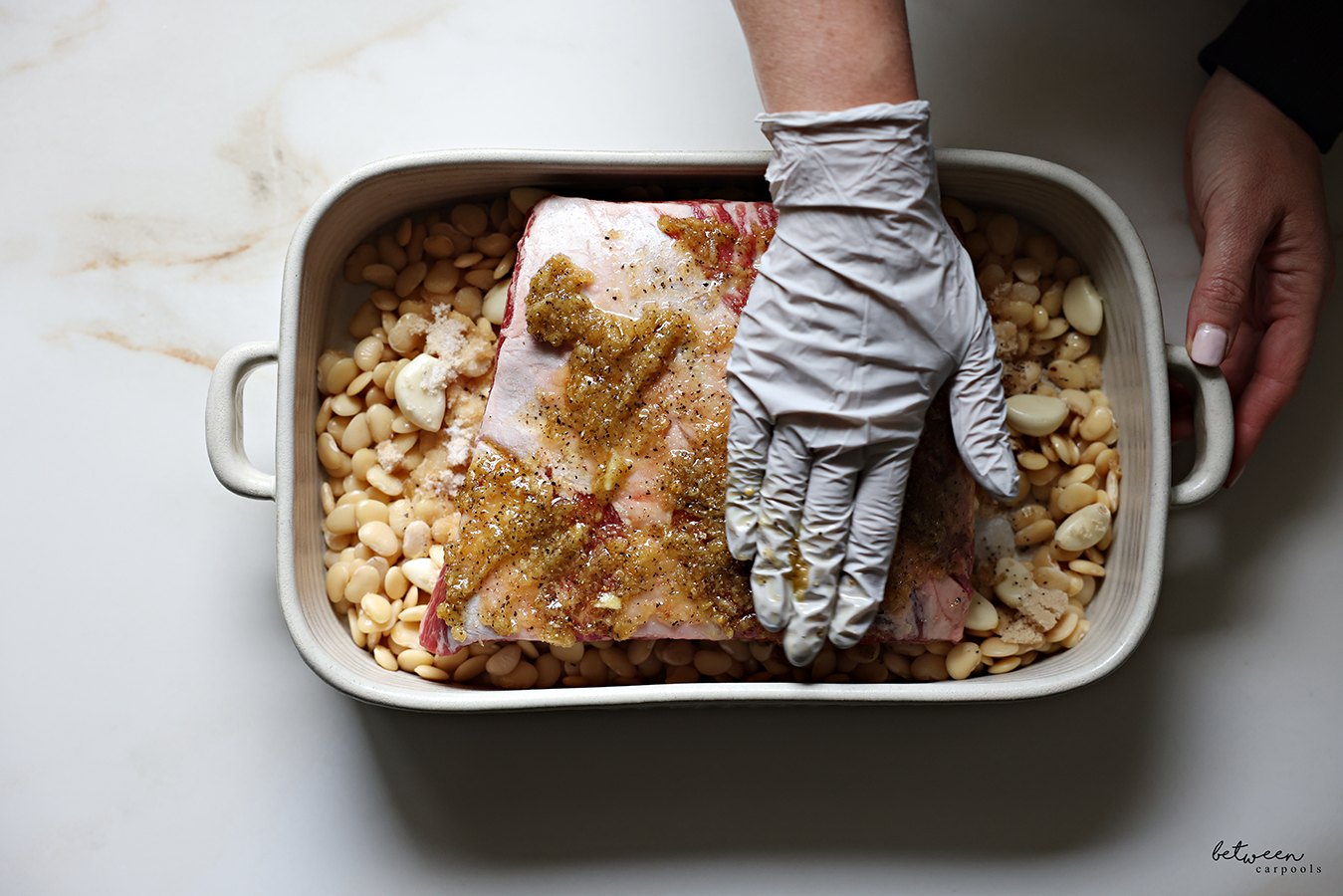 Spreading rub over flanken roast (over lima beans) in a roasting pan, with a gloved hand.