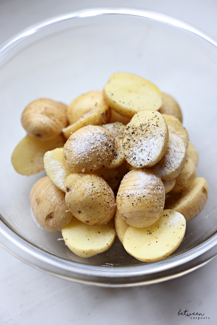Hasselback baby potato halves in a glass bowl with olive oil, salt, and pepper.