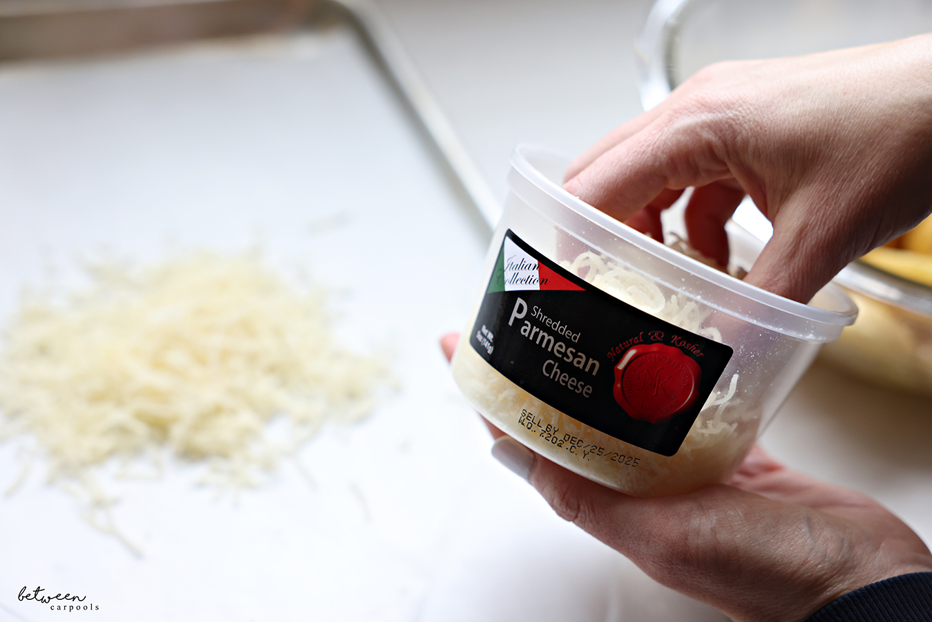 Holding a container of Natural & Kosher Shredded Parmesan Cheese. Some on a parchment lined sheet pan.