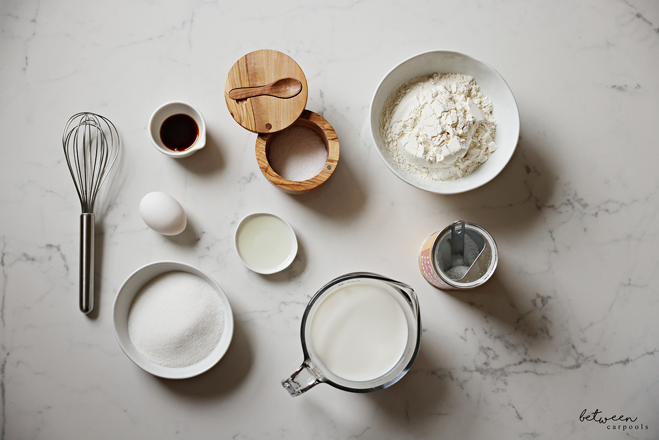 Top down view of pancake ingredients on a countertop: a whisk, an egg, vanilla extract in a condiment bowl, kosher salt in a wood salt cellar, flour in a bowl, an open container of baking powder with a measuring spoon inside, milk in a glass measuring cup, sugar in a bowl and oil on a sauce plate.