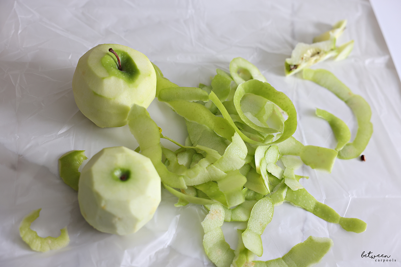 Two peeled Granny Smith apples, with their peels, on a plastic lined kitchen counter.