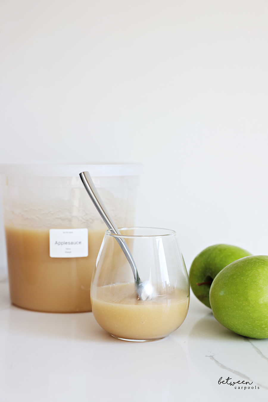 Applesauce in a stemless wine glass with spoon, a labeled deep deli container with applesauce, and 2 Granny Smith apples nearby.