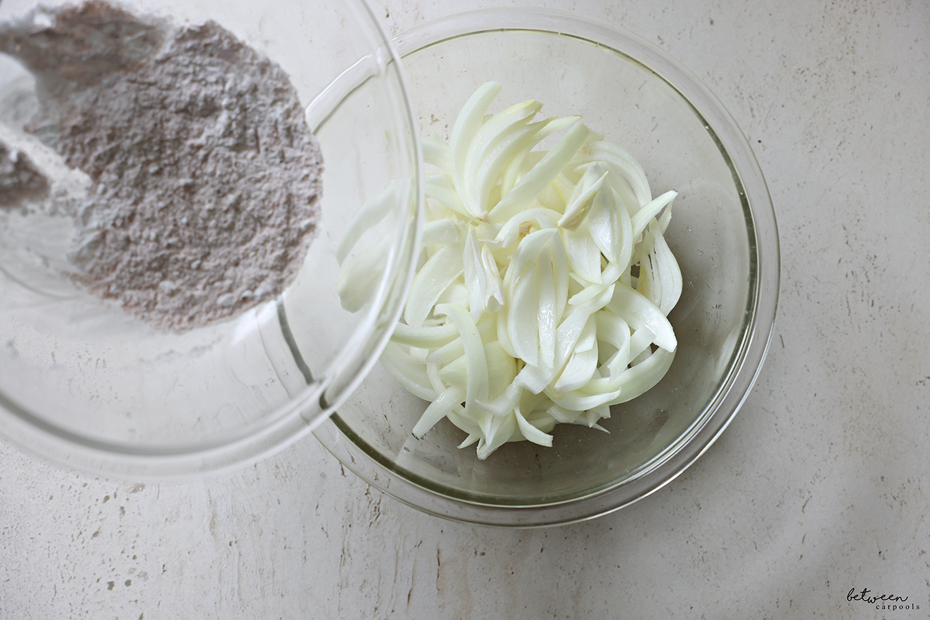 Potato starch and spices from a glass mixing bowl to another bowl with Frenched onions tossed with lemon juice and sugar.