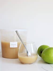 Applesauce in a stemless wine glass with spoon, a labeled deep deli container with applesauce, and 2 Granny Smith apples nearby.